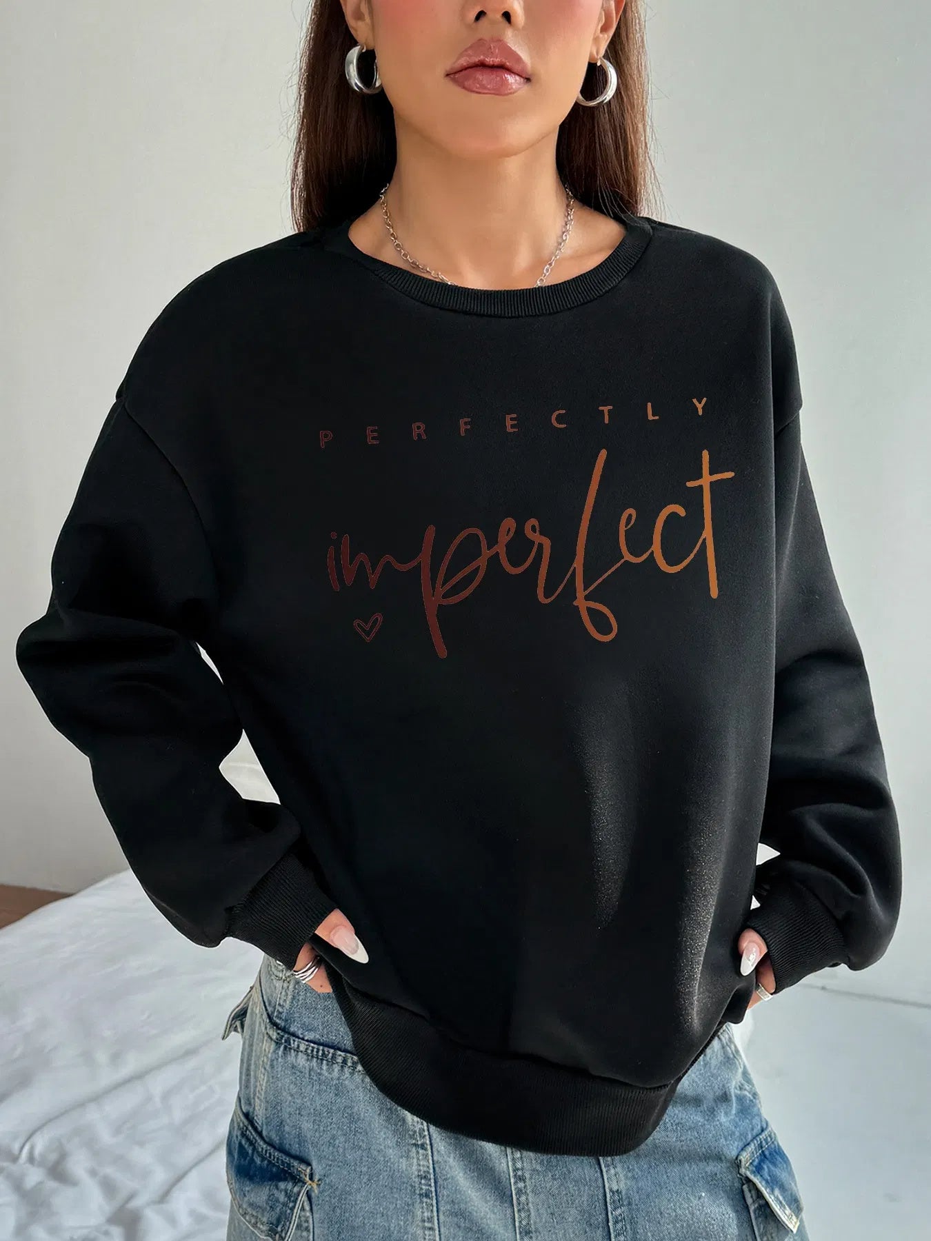 A person wearing a black long-sleeve sweatshirt with the words "perfectly imperfect" printed on the front in a brown/orange script font. They are also wearing a denim skirt and hoop earrings.